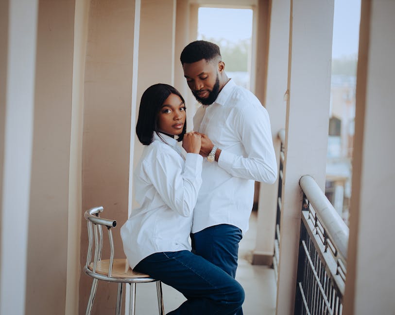 Elegant couple in matching white shirts and jeans sharing a moment on a balcony.