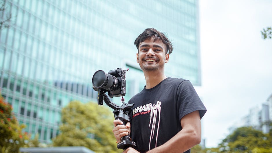 Smiling young filmmaker holding a camera in an urban setting, showcasing his filming equipment.