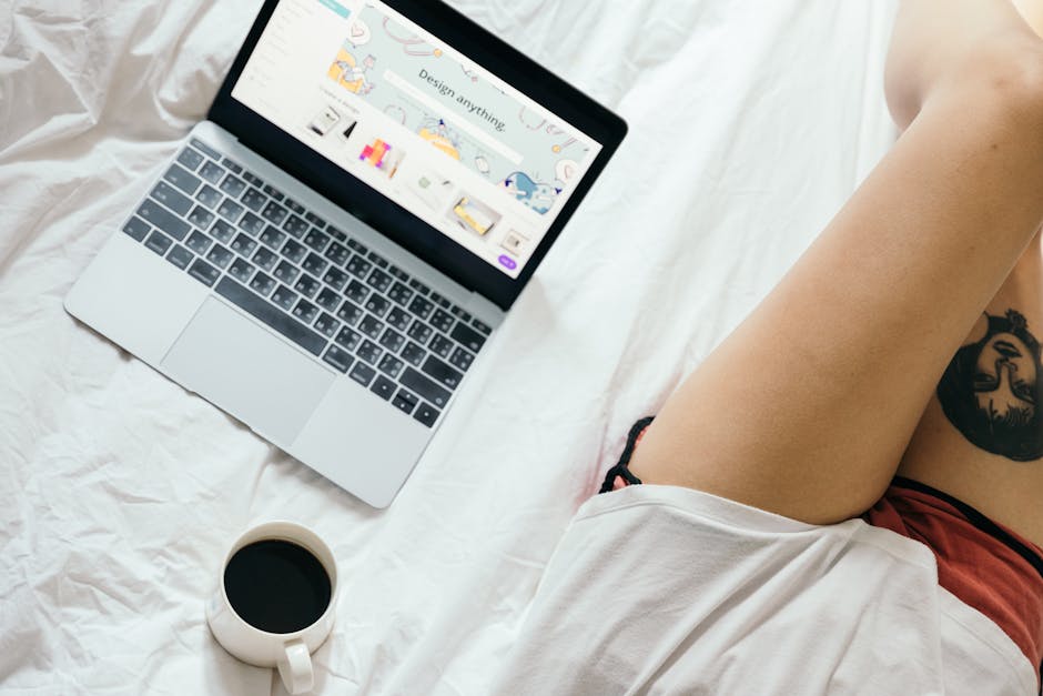 From above of unrecognizable woman with tattoo on leg browsing netbook and drinking coffee while resting on bed in cozy bedroom