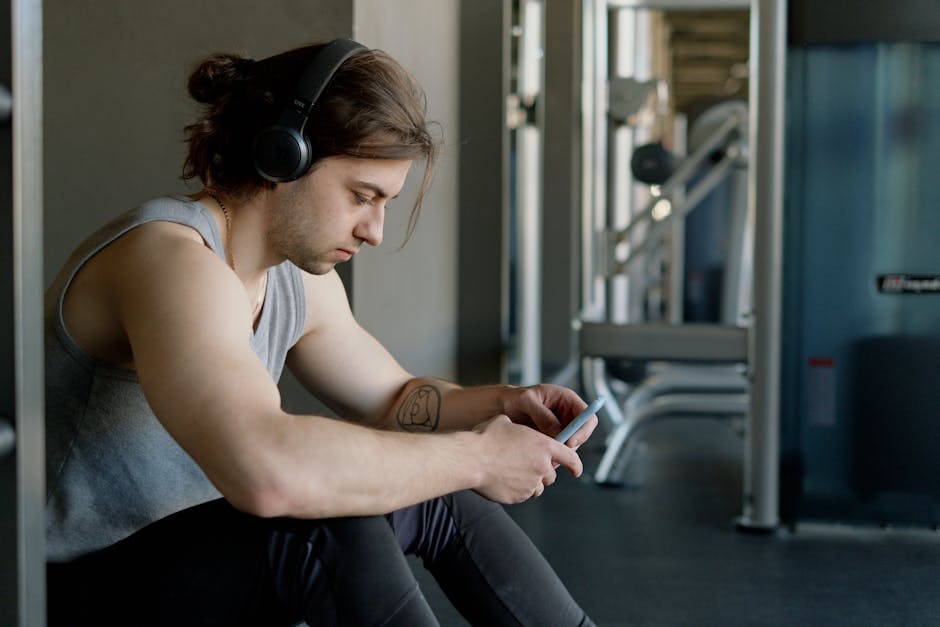 A young man wearing headphones sits in a gym, focused on texting with his smartphone.