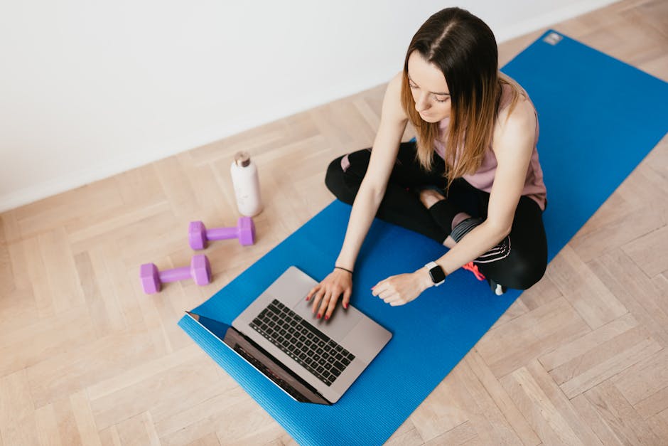 From above of focused young female in activewear sitting on blue yoga mat and browsing laptop