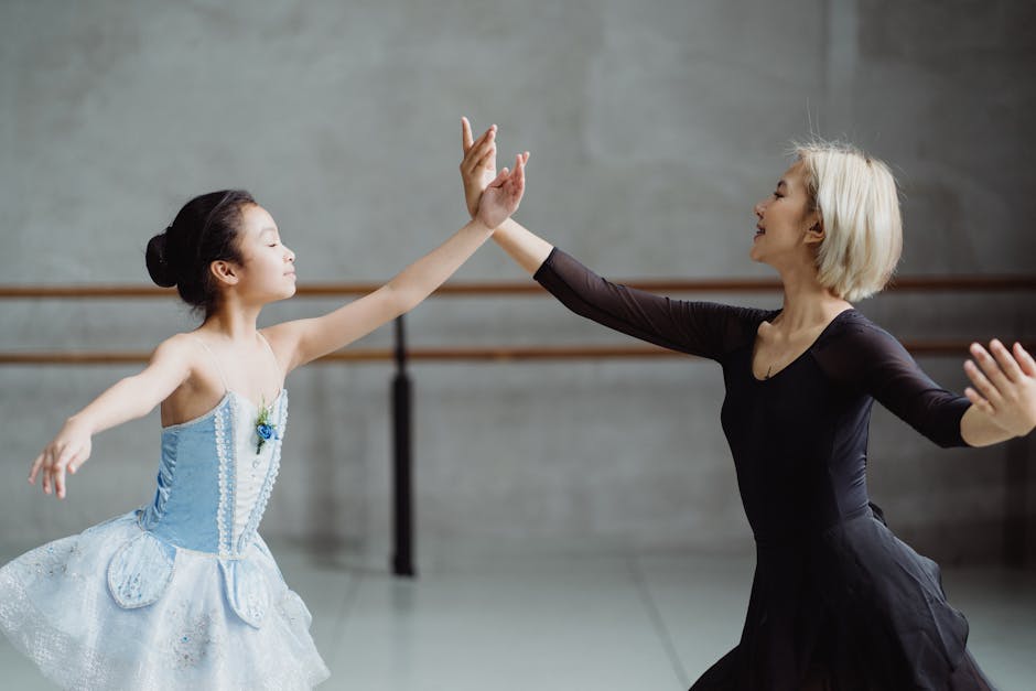 Side view of graceful Asian girl wearing tutu standing near teacher in black outfit with raised arms in ballet studio