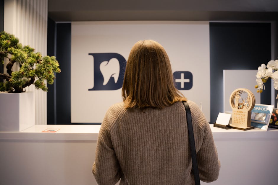 A woman stands at the dental clinic reception wearing a brown sweater, facing away.