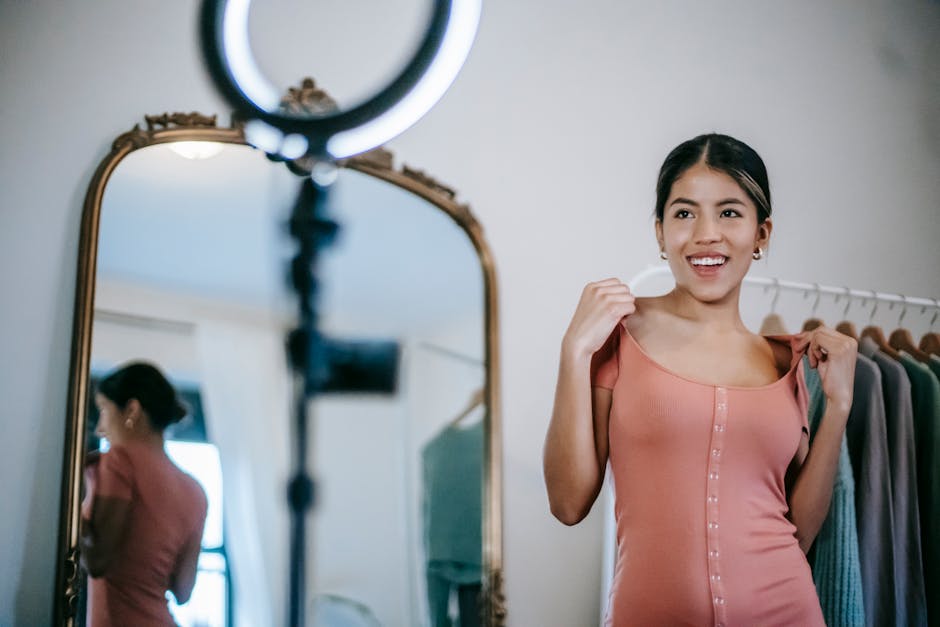 Smiling young ethnic female blogger standing in room and demonstrating pink dress while shooting vlog on smartphone with ring lamp near mirror and wardrobe