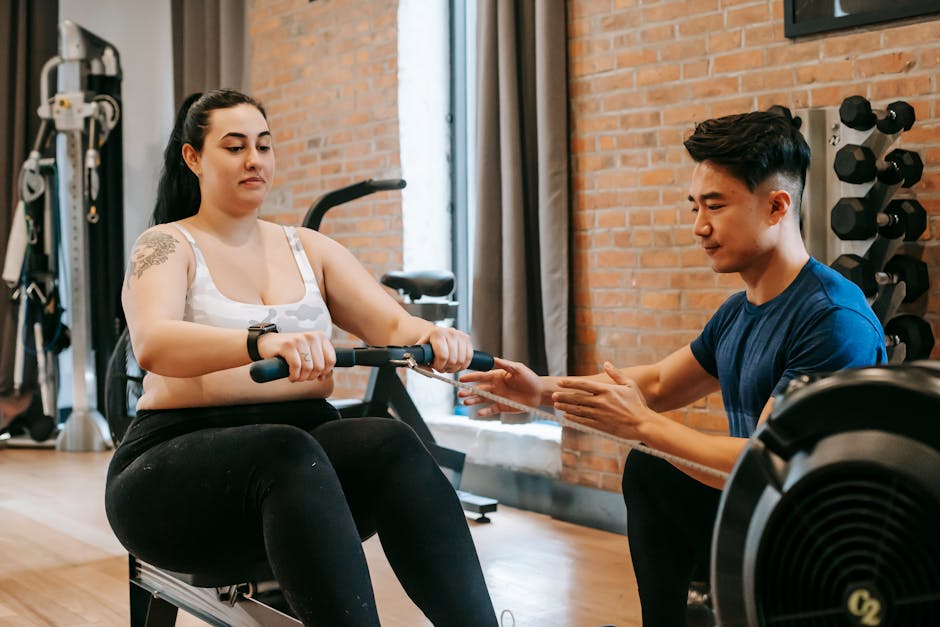 Positive young obese tattooed female in activewear sitting on rowing machine and doing pulling exercise during intense training with Asian male personal coach in modern gym