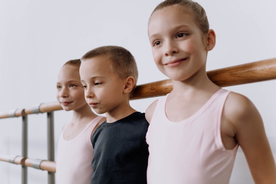 Three children smiling and leaning on a ballet barre in a studio setting.