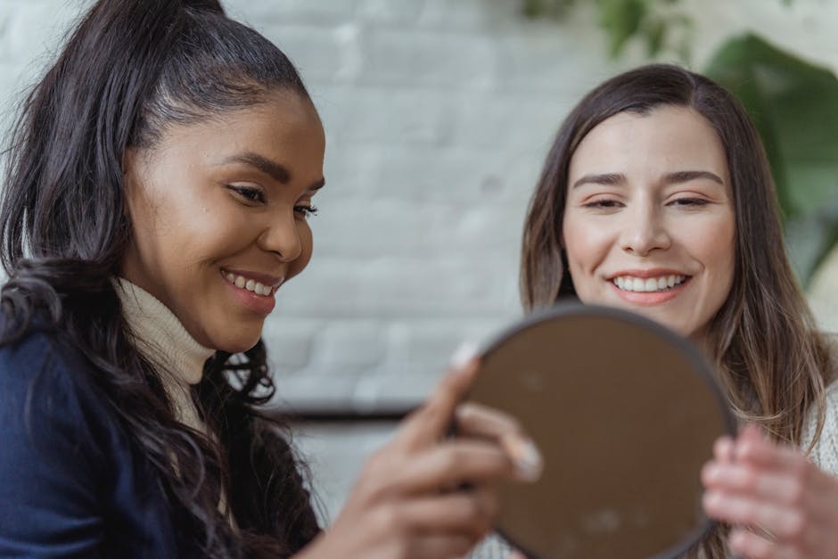 Happy young black female visagiste with long dark hair holding round mirror while showing applied makeup to smiling client in light studio