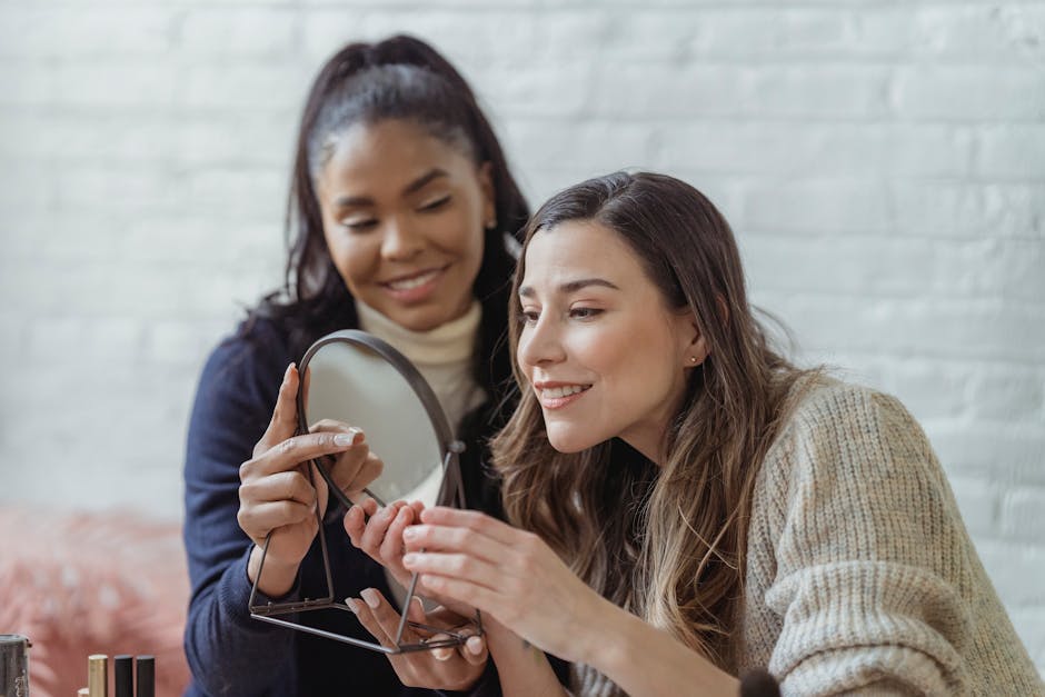 Delighted young woman smiling and looking in round mirror after applying makeup by African American female visagiste in beauty salon