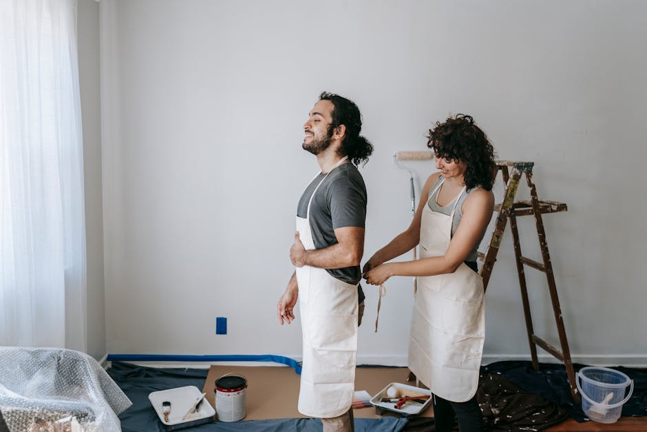 Side view of woman tying apron to positive ethnic man while preparing for painting wall in light room with wooden ladder