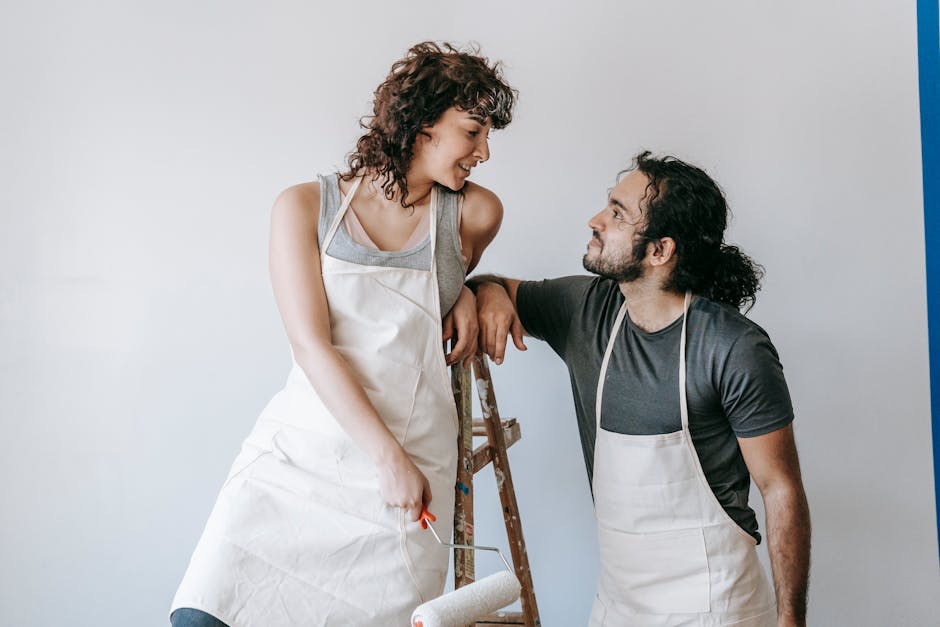 Smiling couple in aprons painting a wall with a roller, enjoying the renovation process.