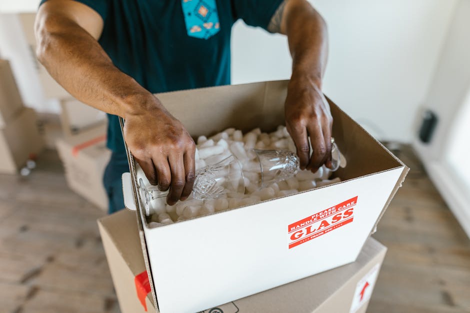 Hands carefully packing glassware into a cardboard box with padding for safe transport.