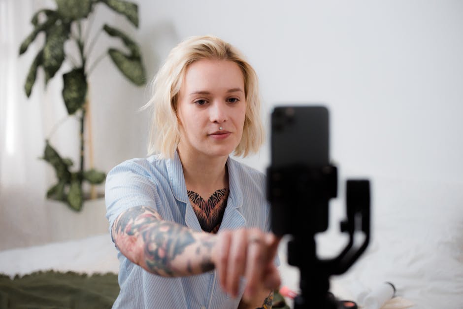 Tattooed woman with blonde hair adjusts smartphone on tripod for vlog setup indoors.