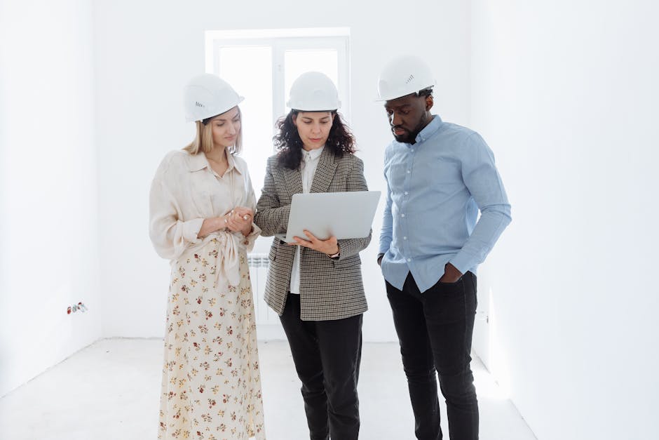 Three professionals in hard hats examining floor plans on a laptop indoors.