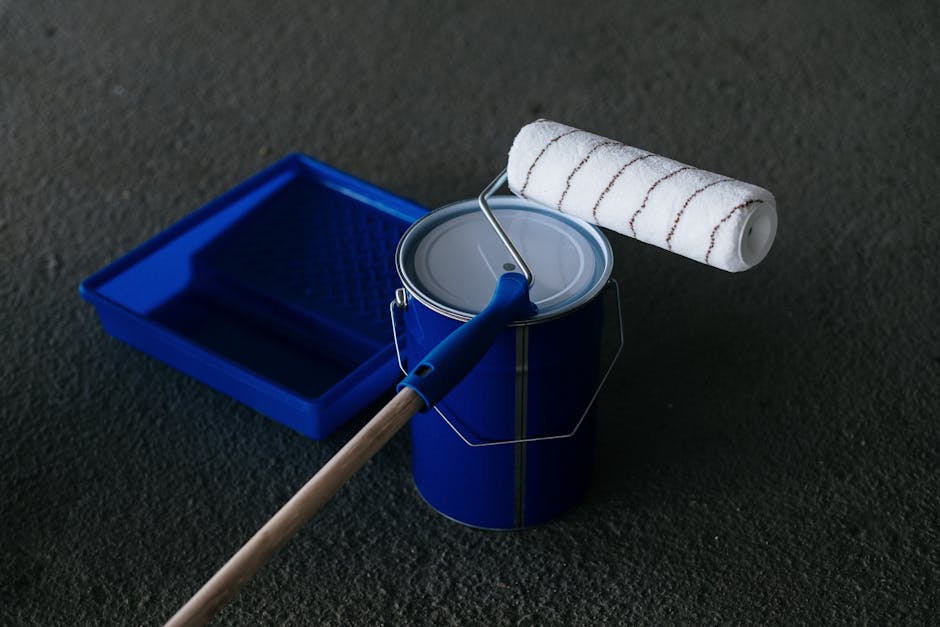 Close-up of a paint can, roller, and tray on a textured floor, perfect for DIY projects.