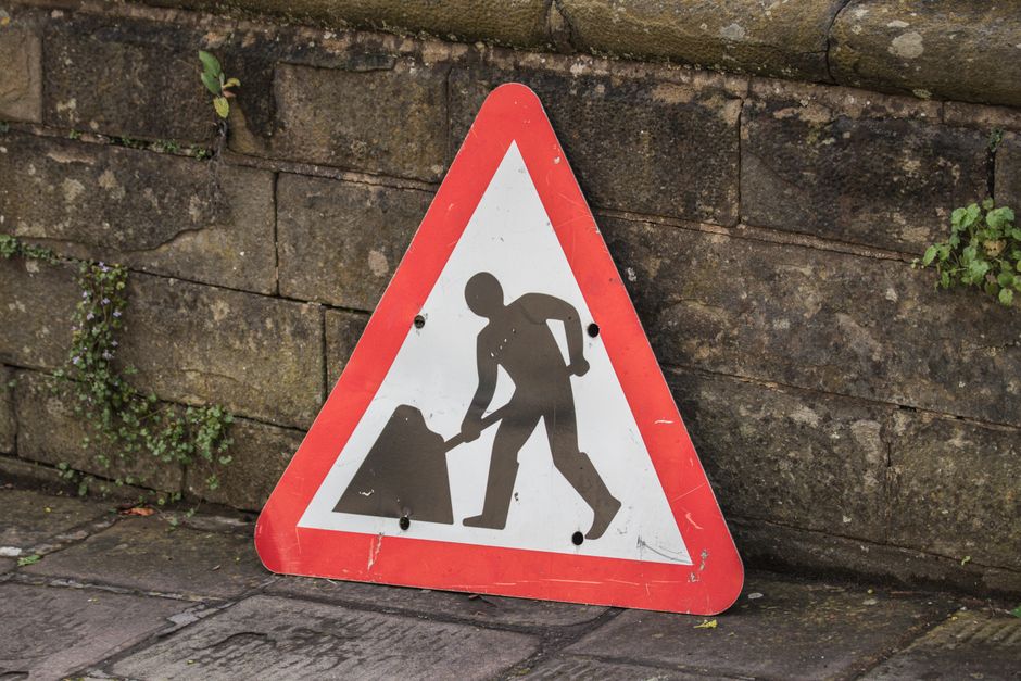 Triangular road work sign placed on a city sidewalk beside a stone wall.