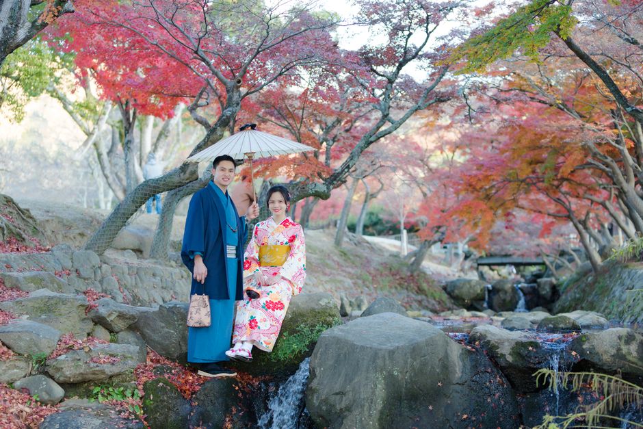 A couple dressed in traditional attire enjoying a scenic autumn landscape with vibrant trees and a flowing stream.