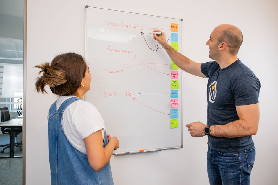 Two colleagues brainstorm marketing strategies on a whiteboard in a modern office setting.