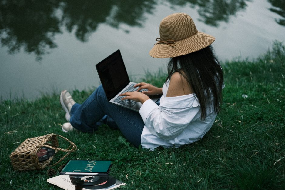 Woman wearing a hat using a laptop while sitting by a tranquil lake in a park.