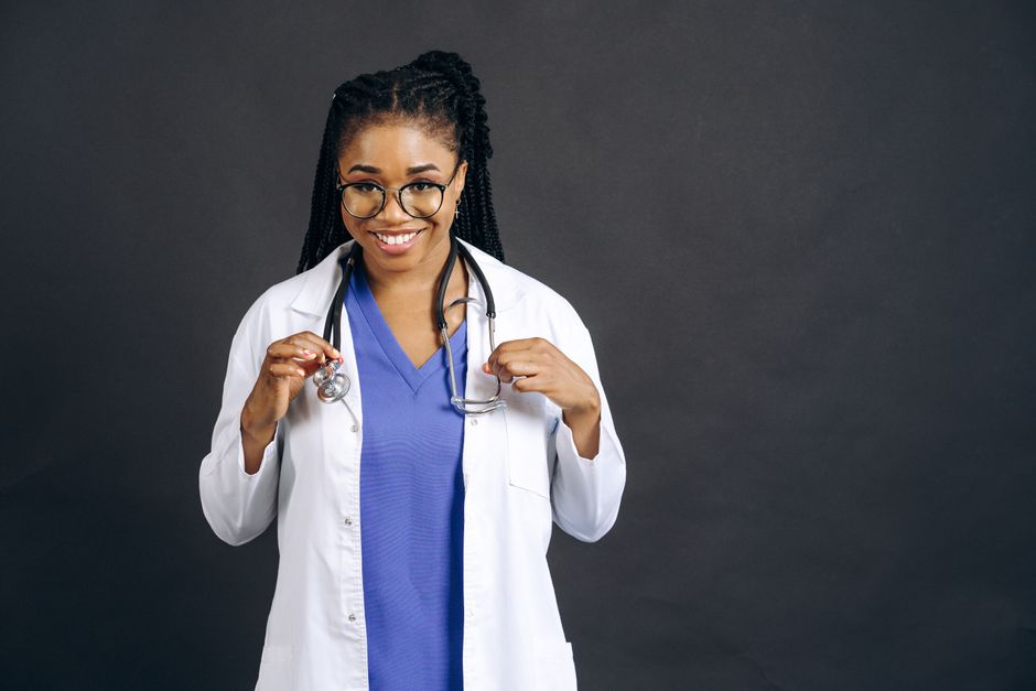 Portrait of a smiling female doctor wearing eyeglasses and a lab coat against a gray background.