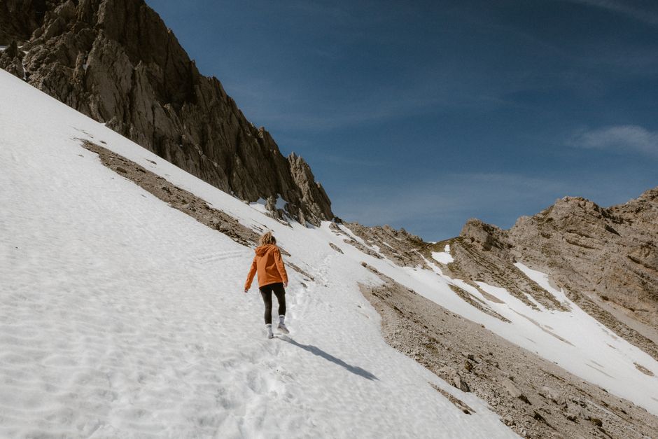A lone hiker in orange ascends a snowy mountain under a clear blue sky.