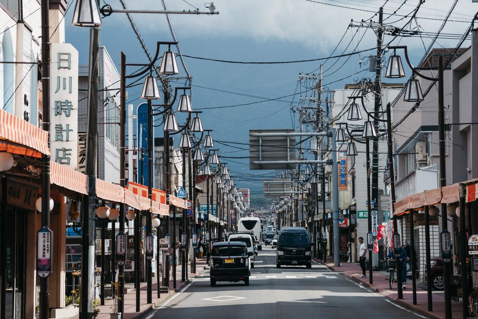 A lively Japanese street with shops, traffic, and mountain background.