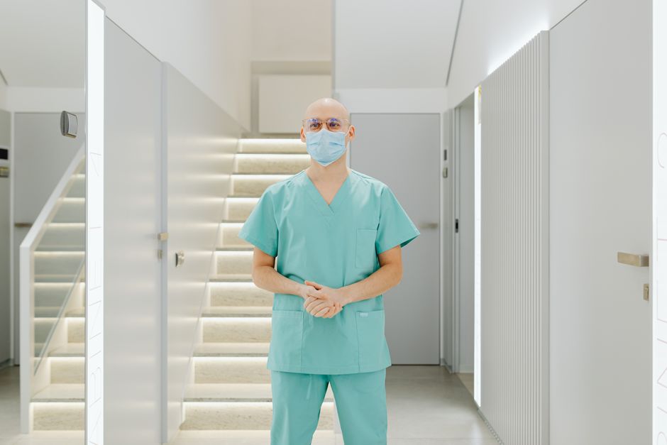 Healthcare worker in scrub suit standing indoors with face mask in clinic hallway.