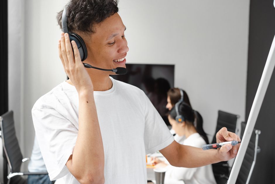 A cheerful customer service agent with headphones writing on a whiteboard in an office setting.