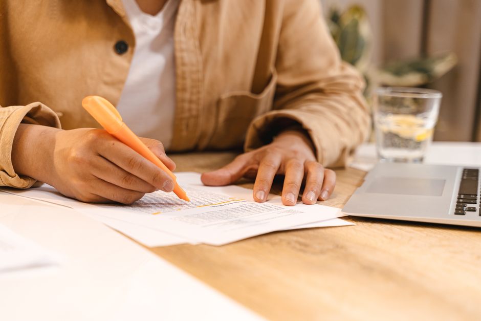 Person highlighting notes at a desk with a laptop and glass of water nearby.